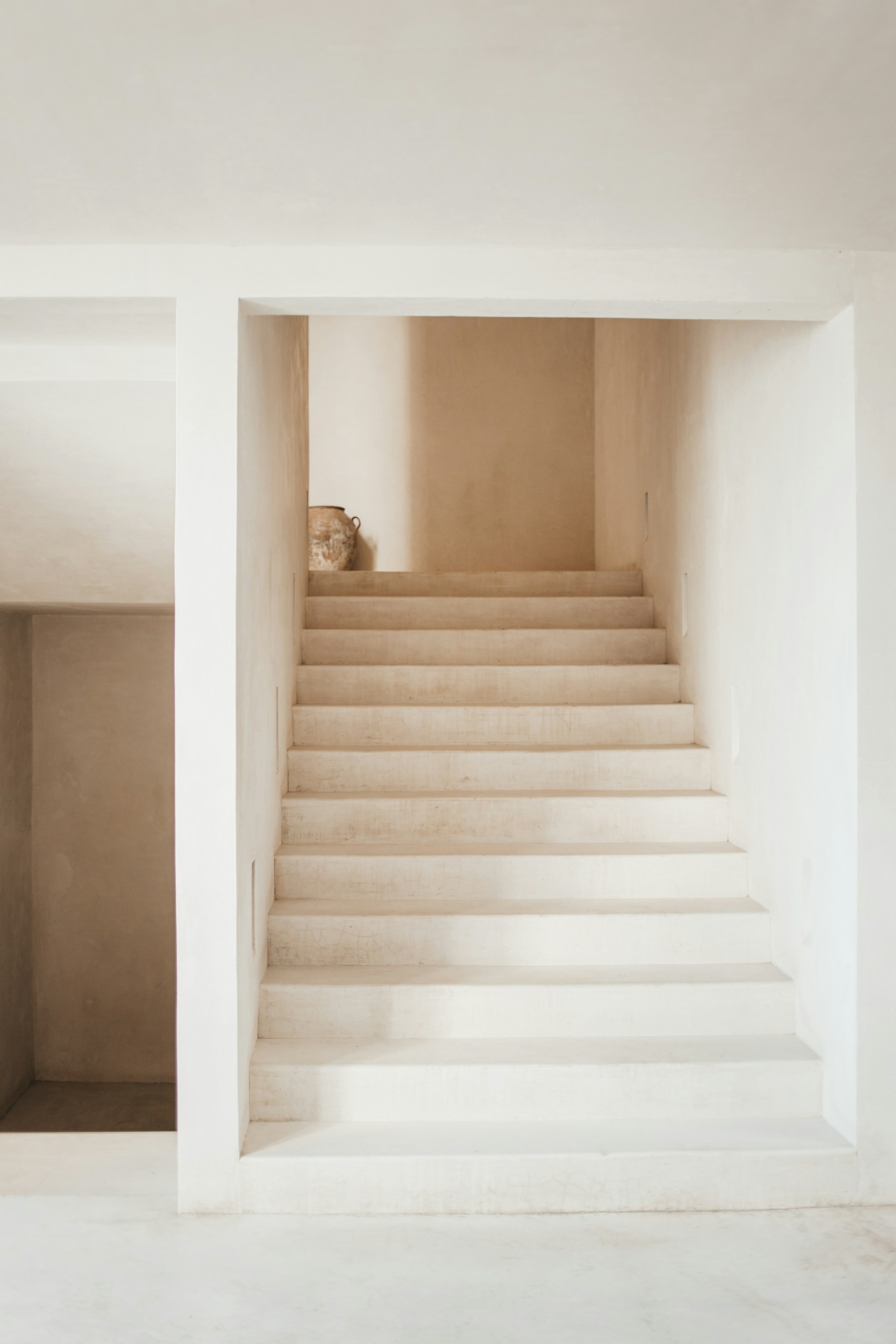 Lime-plaster staircase rising to a soft-lit landing, a single terracotta vessel resting at the top